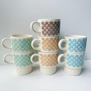 May include: A set of six ceramic mugs arranged in three stacks of two. Each mug features a white base with a repeating checkerboard pattern of small flowers in brown, orange, green, or blue. The lower portion of each mug has vertical ridges.
