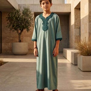 May include: A light sage green thobe featuring long sleeves and a dark green embroidered V-neck collar. The garment reaches the ankles and includes matching dark green trim on the sleeve cuffs, providing a clean and structured appearance.
