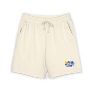 Puede incluir: Pantalones cortos de color blanco roto con cintura elástica, cordón ajustable y dos bolsillos laterales. En la pernera izquierda lleva un gráfico ovalado azul con el texto BE KIND en letras blancas junto a un icono de flor amarilla.