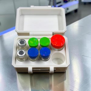 May include: A white plastic storage case containing six small glass vials with colourful caps. Two vials have silver metal caps, two have blue caps with iridescent flecks, two have green caps with iridescent flecks, and one has a large red cap.