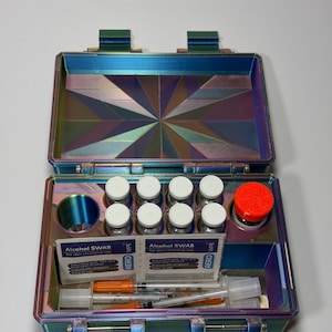 May include: An open, iridescent, multi-colored storage case containing eight small glass vials with white caps, one larger vial with a bright orange cap, two alcohol swab packets labeled "Alcohol SWAB," and two clear plastic syringes.