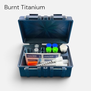 Könnte beinhalten: Ein dunkelblauer Kunststoffkoffer mit der Aufschrift Burnt Titanium, der medizinisches Zubehör enthält. Das Set umfasst zwei durchsichtige Fläschchen, zwei Flaschen mit blauem Deckel, zwei mit grünem Deckel, zwei Alkoholtupfer und zwei Spritzen.