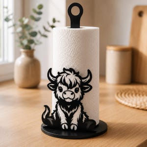 Pode incluir: Um suporte para papel toalha em metal preto com um recorte decorativo da silhueta de uma vaca Highland na base. O suporte inclui uma base circular e uma haste vertical com uma alça em anel no topo, segurando um rolo de papel toalha branco.
