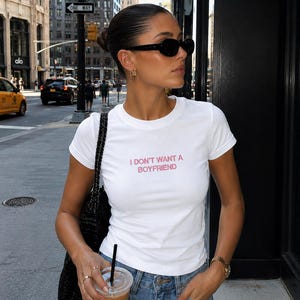 May include: A white short-sleeved crew neck t-shirt featuring the text I DON'T WANT A BOYFRIEND printed in pink letters on the chest. The shirt is paired with blue denim jeans and a black textured shoulder bag with a chain strap.