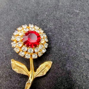 May include: A gold-tone flower brooch featuring a large, round, faceted red center stone surrounded by a halo of small, clear, sparkling rhinestones. The stem and two textured leaves are crafted from polished gold-tone metal, completing the floral design.