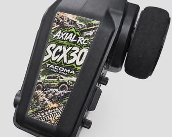 May include: A black radio control transmitter featuring a rectangular sticker with the text Axial RC SCX30 Tacoma TRD PRO and a graphic of a green pickup truck driving over rocky terrain. The device includes a black steering wheel on the side.