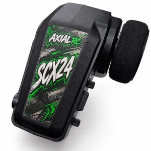 May include: A black radio control transmitter featuring a green and white graphic panel with the text Axial RC and SCX24. The device includes a side-mounted black steering wheel, control knobs, and a textured grip surface for remote operation.