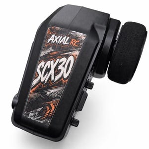 May include: A black radio control transmitter featuring a foam-covered steering wheel on the side. The front panel displays a graphic sticker with the text AXIAL RC and SCX30 in white and orange lettering over a dark, textured background.