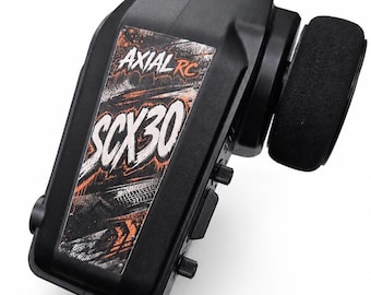 May include: A black radio control transmitter featuring a foam-covered steering wheel on the side. The front panel displays a graphic sticker with the text AXIAL RC and SCX30 in white and orange lettering over a dark, textured background.