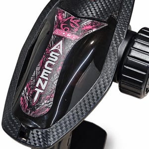 May include: A black remote control transmitter for a radio-controlled vehicle featuring a carbon fiber pattern finish. The center panel displays the text REDCAT ASCENT in white letters over a pink and black graphic design with tire tread patterns.