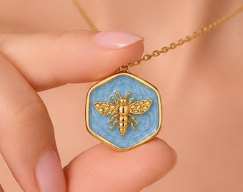 Puede incluir: Un collar de tono dorado con un colgante hexagonal que presenta un fondo de esmalte azul claro y un emblema de abeja dorado, detallado y en relieve, en el centro. El colgante cuelga de una delicada cadena dorada con un acabado pulido.