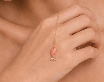 Puede incluir: Una delicada pulsera de cadena dorada con un pequeño dije de pez de color coral y textura marcada. El dije cuelga de una fina cadena dorada que se conecta a un anillo, creando una pieza de joyería para la mano con una estética sutil y orgánica.