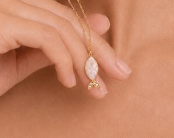 Puede incluir: Un collar de tono dorado con un pequeño colgante en forma de pez. El colgante tiene una incrustación ovalada de color blanco iridiscente para el cuerpo y una sección de cola dorada. El collar cuelga de una cadena de metal fina y delicada.