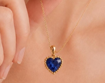 May include: A gold chain necklace featuring a heart-shaped pendant with a faceted deep blue gemstone at the center. The pendant is framed by a delicate beaded gold border, and the entire piece is suspended from a thin, polished gold link chain.