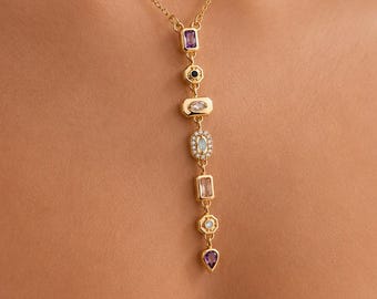 May include: A gold-tone chain necklace featuring a vertical drop pendant composed of seven distinct multi-colored gemstones in various shapes, including rectangular, oval, and pear-cut stones in shades of purple, blue, clear, and soft pink.