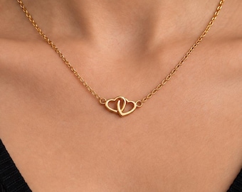 Puede incluir: Un collar de tono dorado con una cadena fina y un colgante central formado por dos contornos de corazón entrelazados. El metal tiene un acabado liso y pulido, y la cadena se conecta directamente a los lados de los corazones.