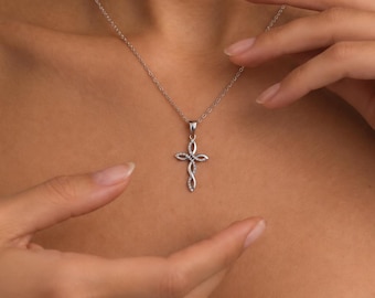 Puede incluir: Un collar de tono plateado que presenta un delicado colgante de cruz con un diseño entrelazado de estilo infinito. El colgante cuelga de una cadena fina a juego, creando una pieza de joyería refinada y sutil.