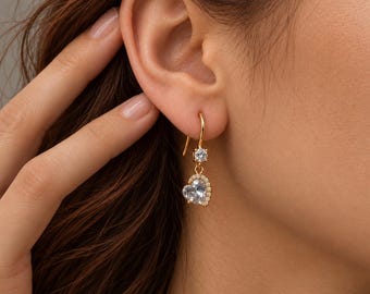 Puede incluir: Un pendiente colgante dorado con una pequeña piedra redonda transparente en la parte superior y un colgante de piedra transparente en forma de corazón. La piedra en forma de corazón está enmarcada por una hilera de pequeñas piedras.
