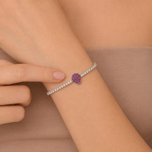 May include: A silver-toned tennis bracelet featuring a row of clear, round-cut stones and a central teardrop-shaped charm encrusted with small, vibrant pink stones. The bracelet is designed with a flexible chain structure and a secure clasp mechanism.