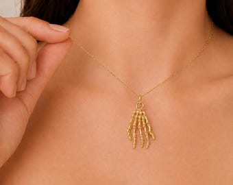 Puede incluir: Un collar de tono dorado con un colgante en forma de mano esquelética. El colgante cuelga de una cadena fina y delicada de tono dorado. El diseño de la mano esquelética muestra los huesos de los dedos y las articulaciones en un acabado dorado.