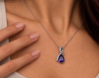 Puede incluir: Un collar de tono plateado con un colgante de amatista morada en forma de lágrima. El colgante presenta un elemento plateado curvado engastado con pequeñas piedras transparentes de corte redondo, suspendido de una cadena fina.
