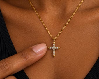 Puede incluir: Un collar de cadena dorada con un colgante de cruz. La cruz está hecha de oro y está engastada con pequeñas piedras redondas y transparentes a lo largo de sus barras verticales y horizontales. El colgante cuelga de una fina cadena de eslabones.