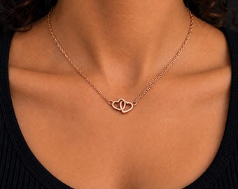 Puede incluir: Un collar de tono oro rosa con una cadena delicada y un colgante compuesto por dos contornos de corazón entrelazados. El metal tiene un acabado liso y pulido, y el colgante se sitúa centrado en la zona de la clavícula del cuello.