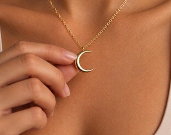 Puede incluir: Un collar de tono dorado con un colgante de media luna pulida suspendido de una delicada cadena de eslabones. El dije de metal liso y curvado refleja la luz, creando un acabado metálico brillante en esta pieza de joyería sencilla.