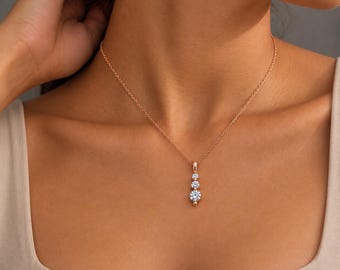 Puede incluir: Un collar de oro rosa con una cadena fina y un colgante vertical engastado con tres piedras redondas, claras y facetadas de diferentes tamaños, dispuestas en orden descendente desde la más pequeña arriba hasta la más grande abajo.
