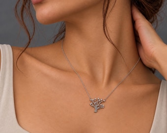 Puede incluir: Un collar de tono plateado con un colgante en forma de árbol estilizado con múltiples ramas, cada una terminando en una pequeña cuenta blanca redonda. El colgante cuelga de una delicada y fina cadena plateada alrededor del cuello.