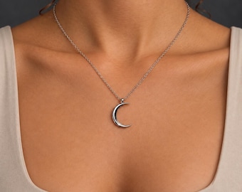 Puede incluir: Un colgante de media luna en tono plateado cuelga de una delicada cadena a juego del mismo tono. El dije de metal liso y pulido tiene forma de luna fina y curva, suspendido en el centro de la cadena sobre una prenda de color neutro.