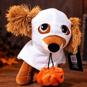May include: A plush dog toy wearing a white ghost costume with a hood, featuring long brown ears, large dark eyes, and a black nose. The stuffed animal holds a small orange pumpkin-shaped treat bucket in its front paws and has a black tag attached.