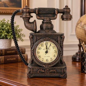 May include: A dark bronze-colored desk clock designed to resemble a vintage telephone. The clock face is circular with a cream background, black Arabic numerals, and the text Old Salem Clock Company. A black coiled cord connects the handset.