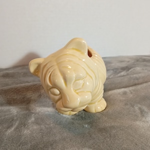 May include: A cream-coloured ceramic tiger figurine with a glossy finish. The tiger is depicted in a crouching position with its head tilted downward, showcasing moulded facial features, ears, and striped patterns across its body.