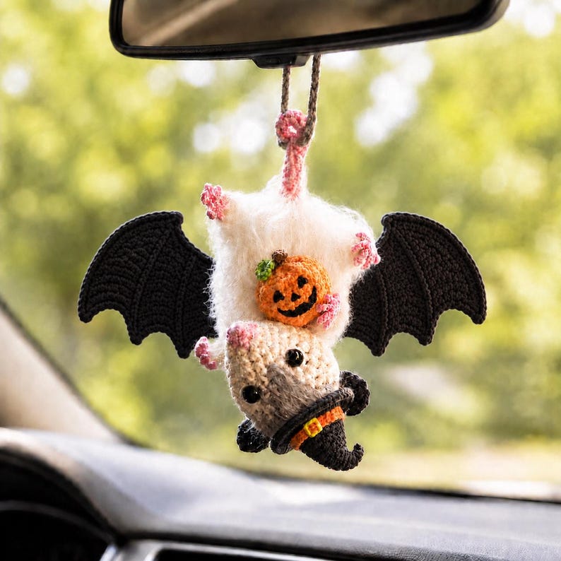 May include: A crocheted hanging ornament featuring a white and tan opossum with large black wings, wearing a black witch hat with an orange band, and holding a small orange jack-o'-lantern pumpkin against its fuzzy white chest.