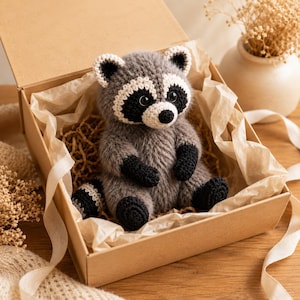 May include: A crocheted raccoon plush toy with grey fur, a white muzzle, and black eye patches. The raccoon has black paws, black feet, and a striped tail. It is nestled inside a square cardboard gift box lined with light-colored tissue paper.