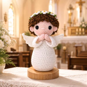 May include: A crocheted angel doll with brown curly hair, a white daisy flower crown, and large black eyes. The figure has white wings, a white dress, and hands pressed together in prayer, standing on a round light-colored wooden base.