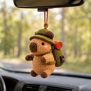 May include: A crocheted capybara hanging ornament featuring a tan body with dark brown paws and nose, wearing a green hat. The figure carries a green backpack with an attached orange rolled sleeping bag, suspended by a brown braided cord.
