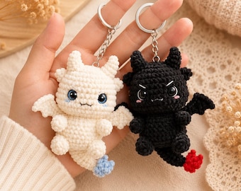 May include: Two crochet dragon keychains, one white with blue-tipped tail and one black with red-tipped tail. Both feature small wings, horns, and large dark eyes. The white dragon has blue eyes, while the black dragon has black eyes and small white fangs.