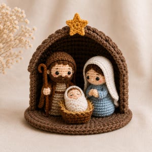May include: A crochet nativity scene featuring a brown arched stable topped with a yellow star. Inside are three figures: a brown-clad figure holding a staff, a blue-clad figure with a white head covering, and a swaddled infant in a brown basket.