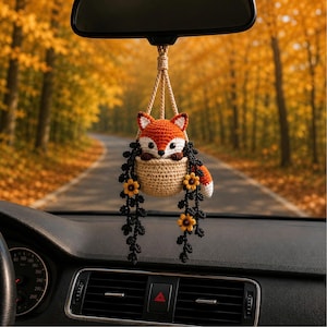 May include: A crocheted fox ornament hanging from a rearview mirror. The fox is orange, white, and black, sitting in a beige basket. Two long, black, trailing vine-like strands with small yellow and brown flowers hang down from the basket.