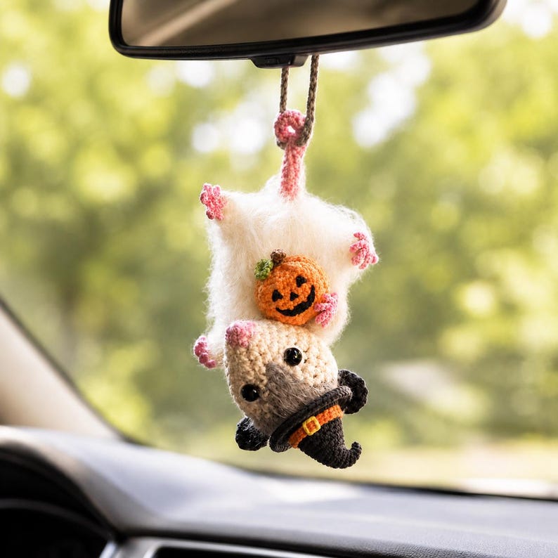 May include: A crocheted hanging ornament featuring a white and beige opossum with pink paws and tail, wearing a black witch hat with an orange band. The opossum is holding a small orange crocheted pumpkin with a green stem and black face.