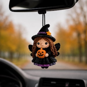 Könnte beinhalten: Ein gehäkelter Hängeanhänger in Form einer kleinen Figur mit braunen Haaren, die einen schwarzen Hexenhut mit gelbem Stern, ein schwarzes Kleid mit violettem Saum und schwarze Flügel trägt und einen kleinen orangefarbenen Kürbis hält.