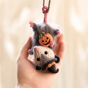 May include: A crocheted hanging ornament featuring a fuzzy gray opossum with a pink nose and feet. The opossum is holding a small orange jack-o'-lantern pumpkin and wearing a black pointed witch hat with an orange band and a yellow buckle.