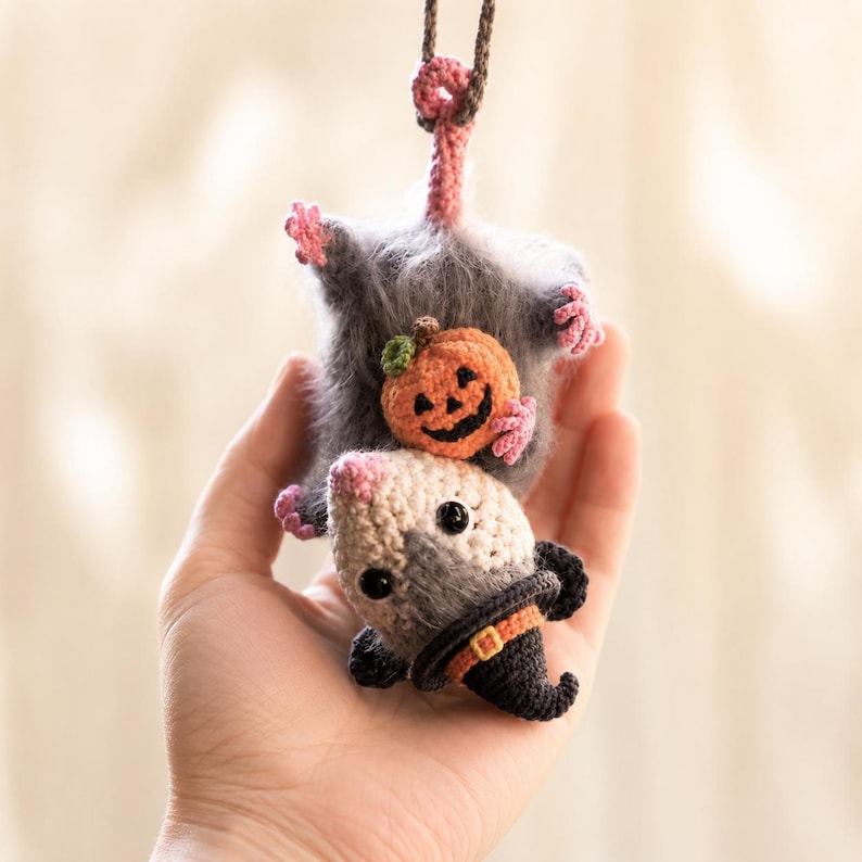 May include: A crocheted hanging ornament featuring a fuzzy gray opossum with a pink nose and feet. The opossum is holding a small orange jack-o'-lantern pumpkin and wearing a black pointed witch hat with an orange band and a yellow buckle.