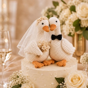 May include: Two crocheted geese cake toppers in cream yarn with orange beaks and feet. One goose wears a white tulle veil and a floral crown, holding a small dried flower bouquet. The other goose wears a small black bow tie. Both sit on a cake.
