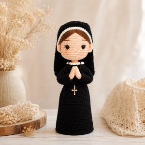 May include: A crocheted doll depicting a figure in a black habit with a white trim, brown hair, and a light skin tone. The doll has hands clasped in a prayer position and wears a small silver-coloured cross pendant hanging from a black cord.
