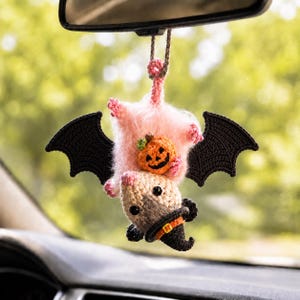 May include: A crocheted hanging ornament featuring a light pink fuzzy creature with large black crocheted wings, holding a small orange jack-o'-lantern. The creature has a beige face, black eyes, and wears a small black witch hat with an orange band.