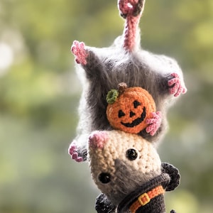 May include: A crocheted opossum hanging by its tail, featuring fuzzy gray fur, pink paws, and a cream-colored face. The opossum holds a small orange crocheted pumpkin and wears a black pointed witch hat with an orange band and yellow buckle.
