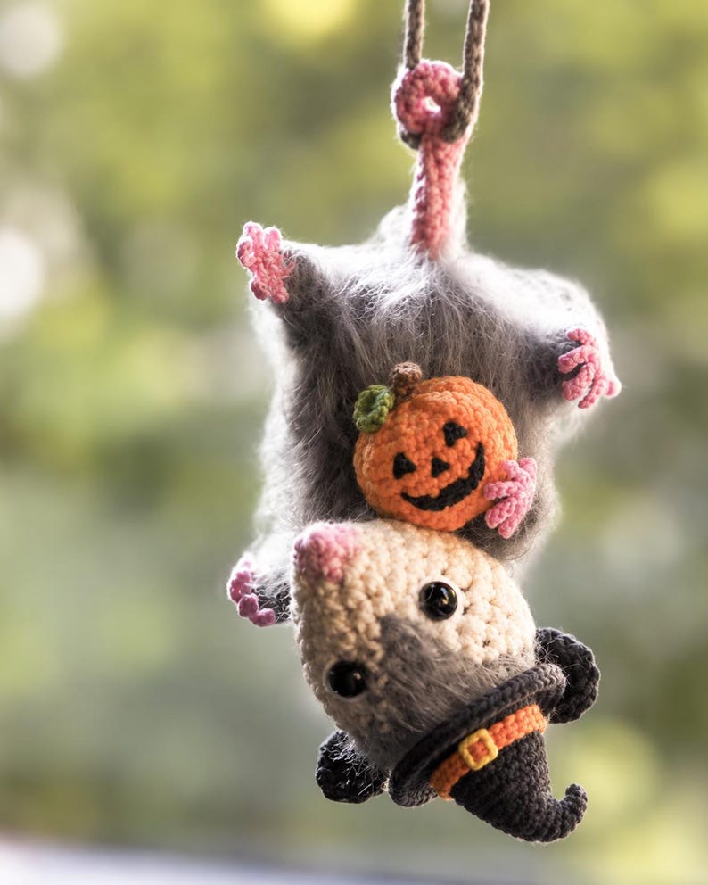 May include: A crocheted opossum hanging by its tail, featuring fuzzy gray fur, pink paws, and a cream-colored face. The opossum holds a small orange crocheted pumpkin and wears a black pointed witch hat with an orange band and yellow buckle.